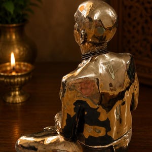 May include: A polished gold-toned metal sculpture of a seated figure viewed from behind. The figure has a smooth, bald head, a beaded necklace, and two distinct U-shaped markings on the upper back and shoulder area, reflecting light.