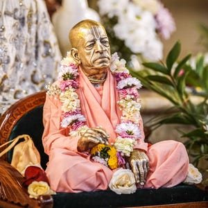 May include: A metallic statue of an elderly person with a shaved head, wearing peach-colored robes and a garland of white, pink, and yellow flowers. The figure is seated in a cross-legged position on a dark-cushioned wooden chair with carved details.