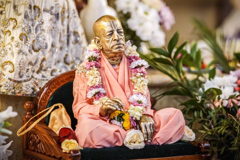 May include: A metallic statue of an elderly person with a shaved head, wearing peach-colored robes and a garland of white, pink, and yellow flowers. The figure is seated in a cross-legged position on a dark-cushioned wooden chair with carved details.