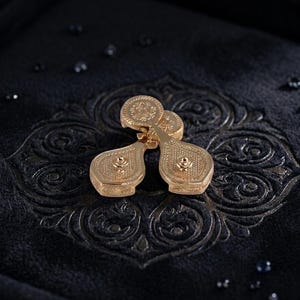 May include: A pair of gold-toned cufflinks featuring a teardrop-shaped design with intricate textured detailing on the surface. Each cufflink has a circular top section and a central raised stud, showcasing a metallic finish and ornate patterns.