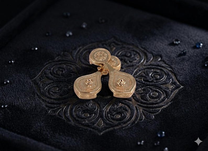 May include: A pair of gold-toned cufflinks featuring a teardrop-shaped design with intricate textured detailing on the surface. Each cufflink has a circular top section and a central raised stud, showcasing a metallic finish and ornate patterns.