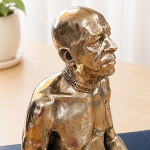 May include: A metallic bronze-toned sculpture of a bald person with a smooth, reflective surface. The figure is depicted from the chest up, featuring a prominent brow, a defined nose, and a layered necklace around the neck, captured in profile.
