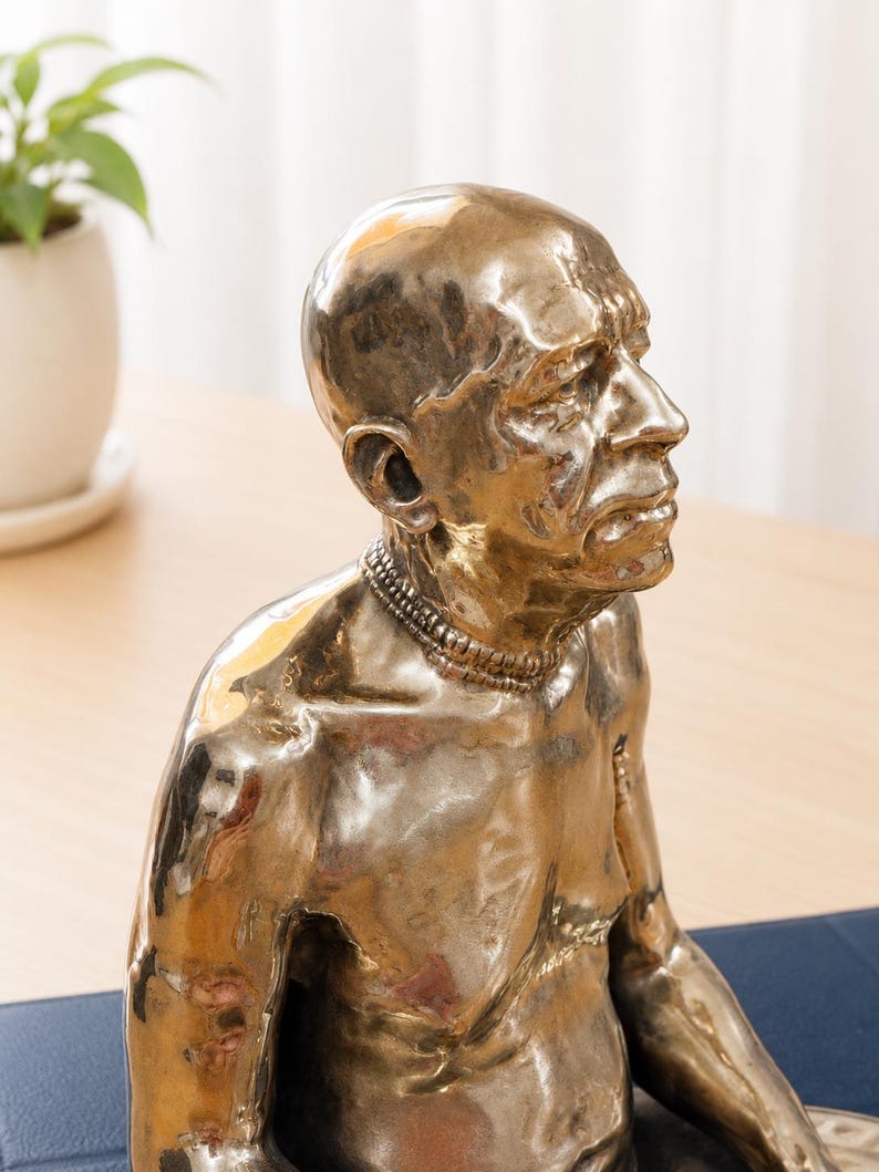 May include: A metallic bronze-toned sculpture of a bald person with a smooth, reflective surface. The figure is depicted from the chest up, featuring a prominent brow, a defined nose, and a layered necklace around the neck, captured in profile.