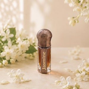May include: A small, hexagonal glass bottle filled with amber-colored liquid, featuring a dark metallic cap and the word Jasmine printed vertically on the front side. The bottle is surrounded by white jasmine flowers and green leaves.