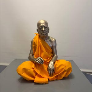 May include: A metallic, silver-toned sculpture of a seated figure with a shaved head, draped in a bright orange fabric robe. The figure is depicted in a meditative pose with hands resting on the legs, featuring realistic facial details and skin texture.