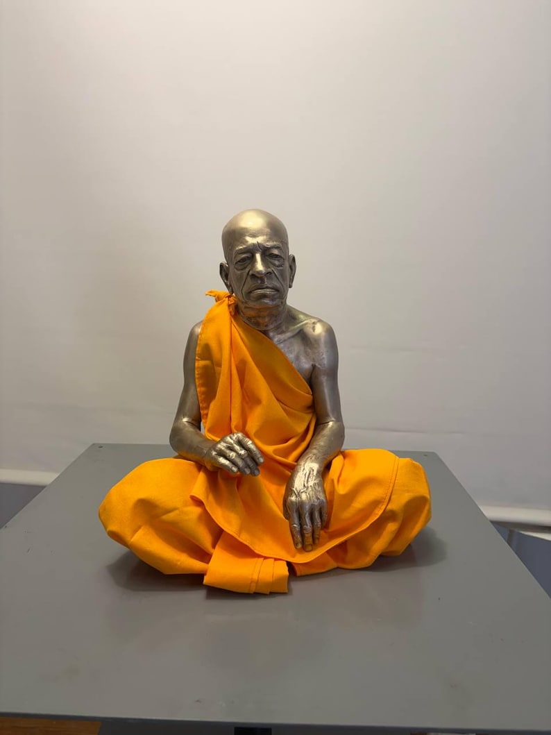 May include: A metallic, silver-toned sculpture of a seated figure with a shaved head, draped in a bright orange fabric robe. The figure is depicted in a meditative pose with hands resting on the legs, featuring realistic facial details and skin texture.