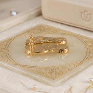 May include: A pair of gold-toned metal flip-flop shaped earrings with intricate engraved detailing, resting on a square, cream-colored stone base featuring gold-colored filigree patterns and surrounded by small white pearls.