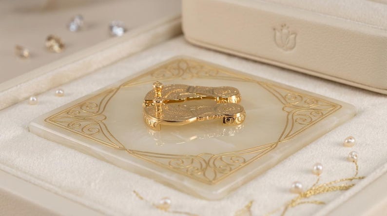 May include: A pair of gold-toned metal flip-flop shaped earrings with intricate engraved detailing, resting on a square, cream-colored stone base featuring gold-colored filigree patterns and surrounded by small white pearls.
