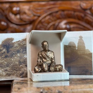 May include: A bronze-colored statue of a seated figure inside a small, open, white box. The box is surrounded by a multi-panel display of sepia-toned photographs. The statue is the focal point, with detailed features. The background includes a wooden surface and carved wooden details.