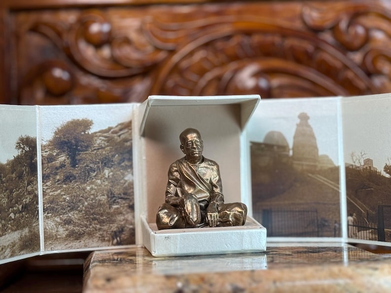 May include: A bronze-colored statue of a seated figure inside a small, open, white box. The box is surrounded by a multi-panel display of sepia-toned photographs. The statue is the focal point, with detailed features. The background includes a wooden surface and carved wooden details.