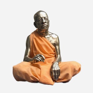 May include: A metallic sculpture of a seated figure with a shaved head, depicted in a meditative pose. The figure has a smooth, reflective silver-toned finish and is draped in a soft, light orange fabric that covers the shoulders and legs.
