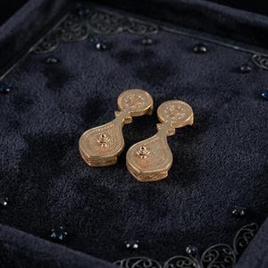 May include: A pair of gold-toned drop earrings featuring a circular top component connected to a larger, pear-shaped base. Both sections exhibit a fine, textured, cross-hatched pattern across their surfaces, with a small central stud detail.