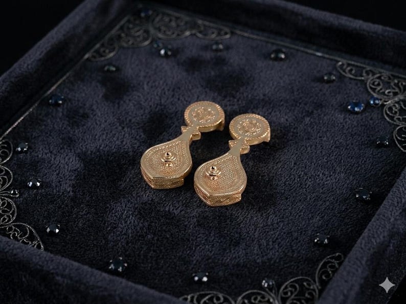 May include: A pair of gold-toned drop earrings featuring a circular top component connected to a larger, pear-shaped base. Both sections exhibit a fine, textured, cross-hatched pattern across their surfaces, with a small central stud detail.