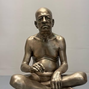 May include: A metallic bronze-toned sculpture of an elderly, bald individual sitting in a cross-legged position. The figure has a serious facial expression, defined musculature, and is depicted without clothing, showcasing detailed skin texture and anatomy.