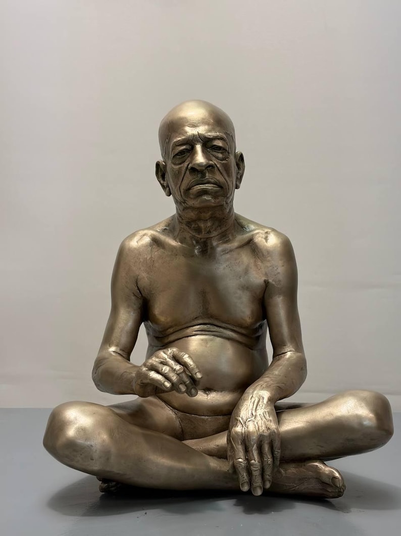May include: A metallic bronze-toned sculpture of an elderly, bald individual sitting in a cross-legged position. The figure has a serious facial expression, defined musculature, and is depicted without clothing, showcasing detailed skin texture and anatomy.
