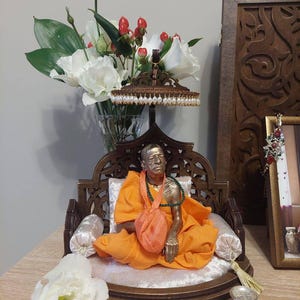 May include: A bronze statue of a religious figure in an orange robe, seated on a decorative wooden chair with a white cushion. The statue is adorned with a green necklace and a small orange bag. A vase of white flowers and red berries is in the background.