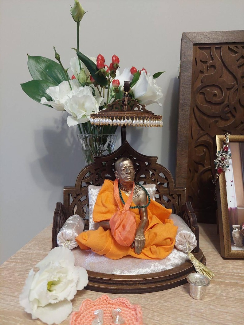 May include: A bronze statue of a religious figure in an orange robe, seated on a decorative wooden chair with a white cushion. The statue is adorned with a green necklace and a small orange bag. A vase of white flowers and red berries is in the background.
