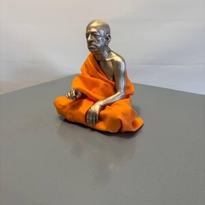 May include: A metallic sculpture of a seated figure with a shaved head, wearing a bright orange fabric robe draped over the shoulders and legs. The figure has a silver-toned finish on the head, chest, and arms, with a necklace visible around the neck.
