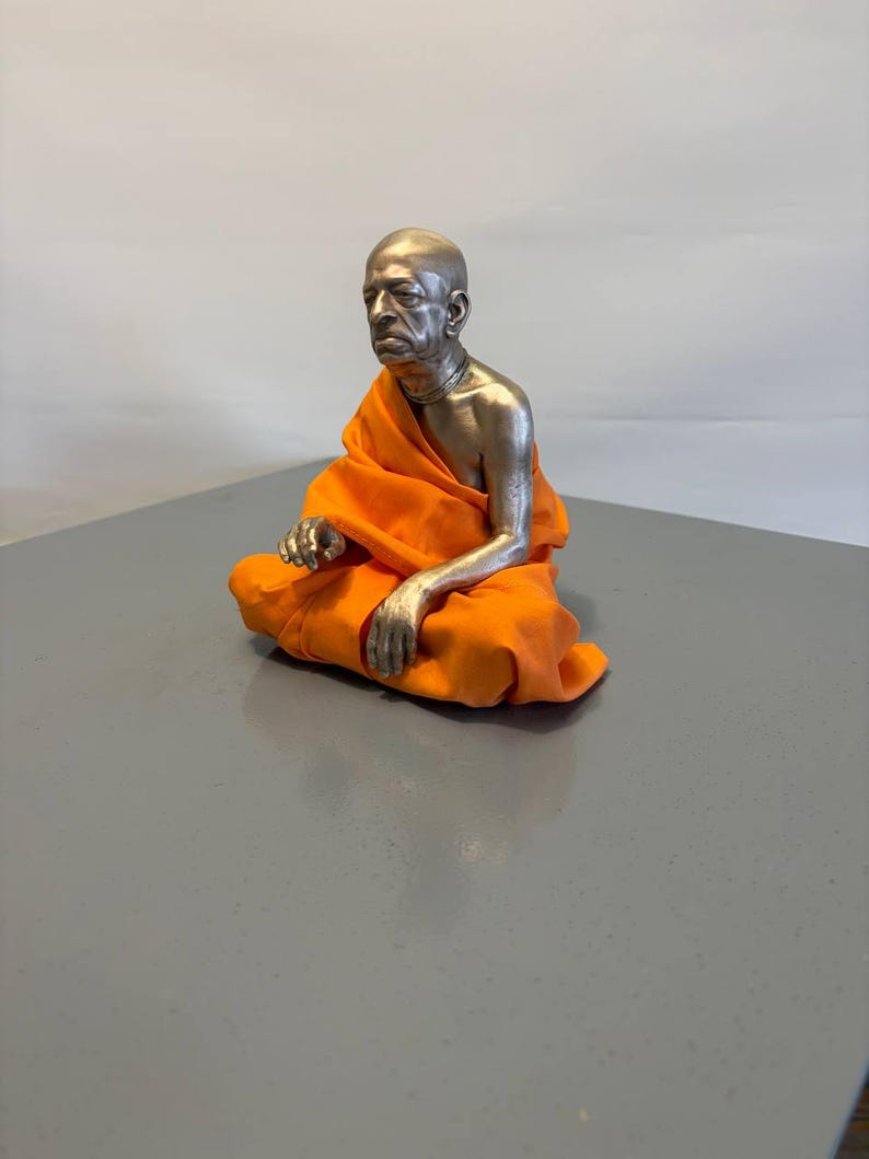 May include: A metallic sculpture of a seated figure with a shaved head, wearing a bright orange fabric robe draped over the shoulders and legs. The figure has a silver-toned finish on the head, chest, and arms, with a necklace visible around the neck.