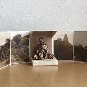 May include: A bronze-colored statue of a seated figure inside a small, open box. The box is surrounded by sepia-toned photographs of landscapes and buildings. The statue is the focal point, with detailed features and a serene expression. The overall theme is religious or spiritual.