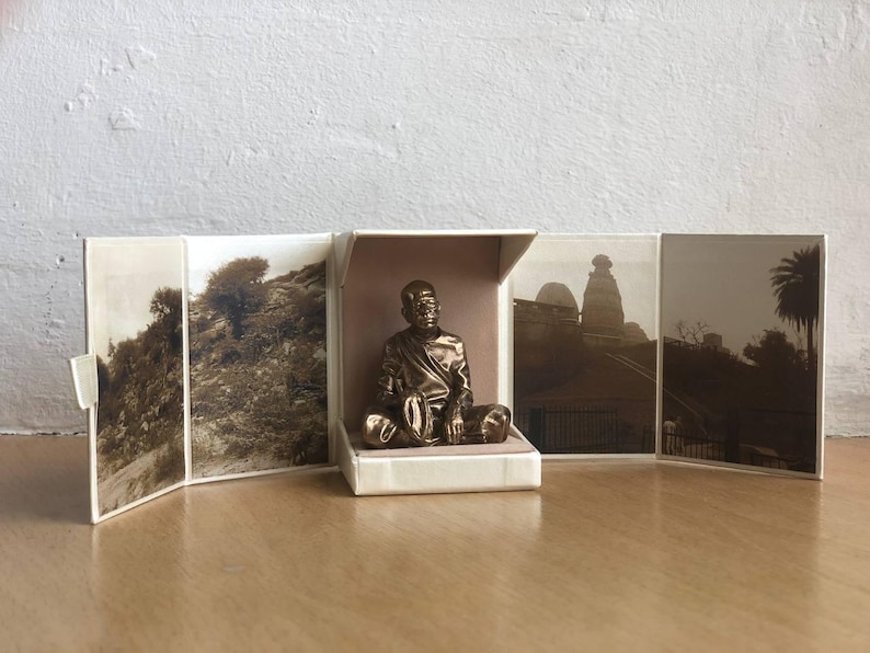 May include: A bronze-colored statue of a seated figure inside a small, open box. The box is surrounded by sepia-toned photographs of landscapes and buildings. The statue is the focal point, with detailed features and a serene expression. The overall theme is religious or spiritual.