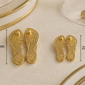 May include: Two pairs of gold-colored metal flip-flop charms with intricate embossed patterns. The larger pair measures 32 mm in length, while the smaller pair measures 22 mm in length. Each charm features a textured sole design and a small post.