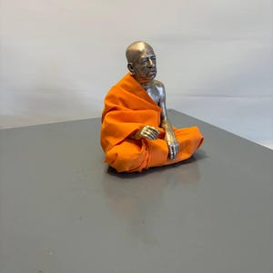 May include: A metallic figurine of a seated person with a shaved head, wearing a bright orange fabric robe draped over one shoulder. The figure has a silver-toned finish on the head, chest, and arms, and is posed in a cross-legged position.
