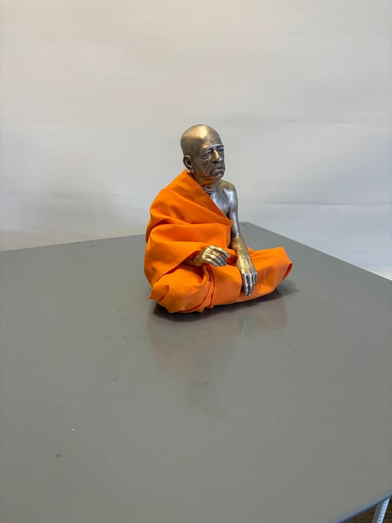 May include: A metallic figurine of a seated person with a shaved head, wearing a bright orange fabric robe draped over one shoulder. The figure has a silver-toned finish on the head, chest, and arms, and is posed in a cross-legged position.