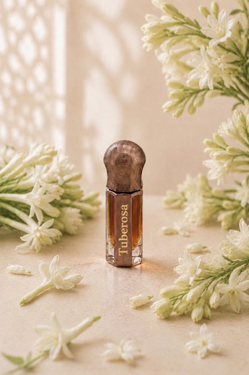 May include: A small, hexagonal glass perfume bottle filled with amber-colored liquid. The bottle features a dark, metallic, rounded cap and the word Tuberosa printed vertically in white lettering on the front face of the glass container.