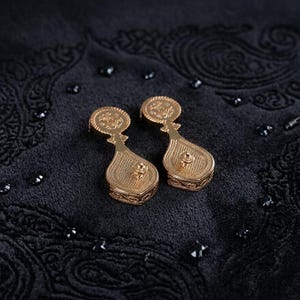 May include: A pair of gold-toned drop earrings featuring a circular top section connected to a larger, teardrop-shaped base. Both components are decorated with intricate, embossed patterns and a central stud detail on the lower portion.