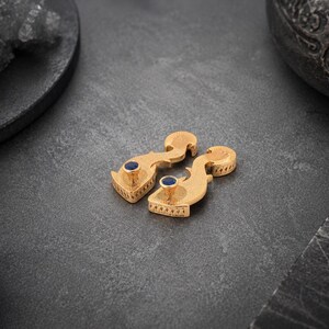 May include: A pair of gold-toned earrings featuring a curved, textured metal body with a small, circular, deep blue stone set at the pointed end. The earrings have a distinct, elongated shape with intricate surface detailing along the edges.