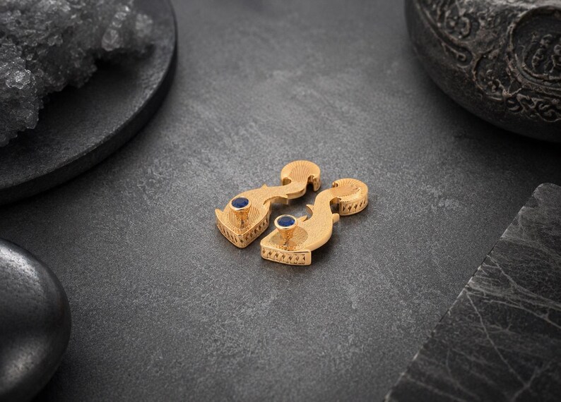 May include: A pair of gold-toned earrings featuring a curved, textured metal body with a small, circular, deep blue stone set at the pointed end. The earrings have a distinct, elongated shape with intricate surface detailing along the edges.