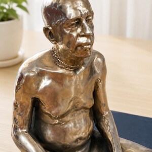 May include: A metallic bronze-toned sculpture of a seated person with a bald head and a textured surface. The figure has a prominent belly, defined facial features, and wears a beaded necklace around the neck, captured in a seated pose.