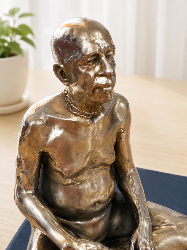 May include: A metallic bronze-toned sculpture of a seated person with a bald head and a textured surface. The figure has a prominent belly, defined facial features, and wears a beaded necklace around the neck, captured in a seated pose.