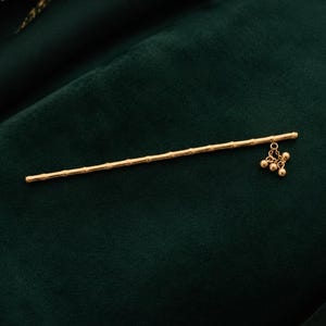 Puede incluir: Un palillo para el cabello de tono dorado con un dise&ntilde;o texturizado similar al bamb&uacute; a lo largo del eje. Un extremo del palillo est&aacute; adornado con un peque&ntilde;o dije colgante que consiste en tres diminutas cuentas redondas doradas en cadenas.