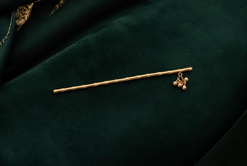 Puede incluir: Un palillo para el cabello de tono dorado con un dise&ntilde;o texturizado similar al bamb&uacute; a lo largo del eje. Un extremo del palillo est&aacute; adornado con un peque&ntilde;o dije colgante que consiste en tres diminutas cuentas redondas doradas en cadenas.