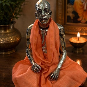 May include: A metallic silver-toned figurine of a seated person with a shaved head, wearing a draped orange fabric garment. The figure features detailed facial expressions and markings on the forehead, presented in a seated, meditative posture.