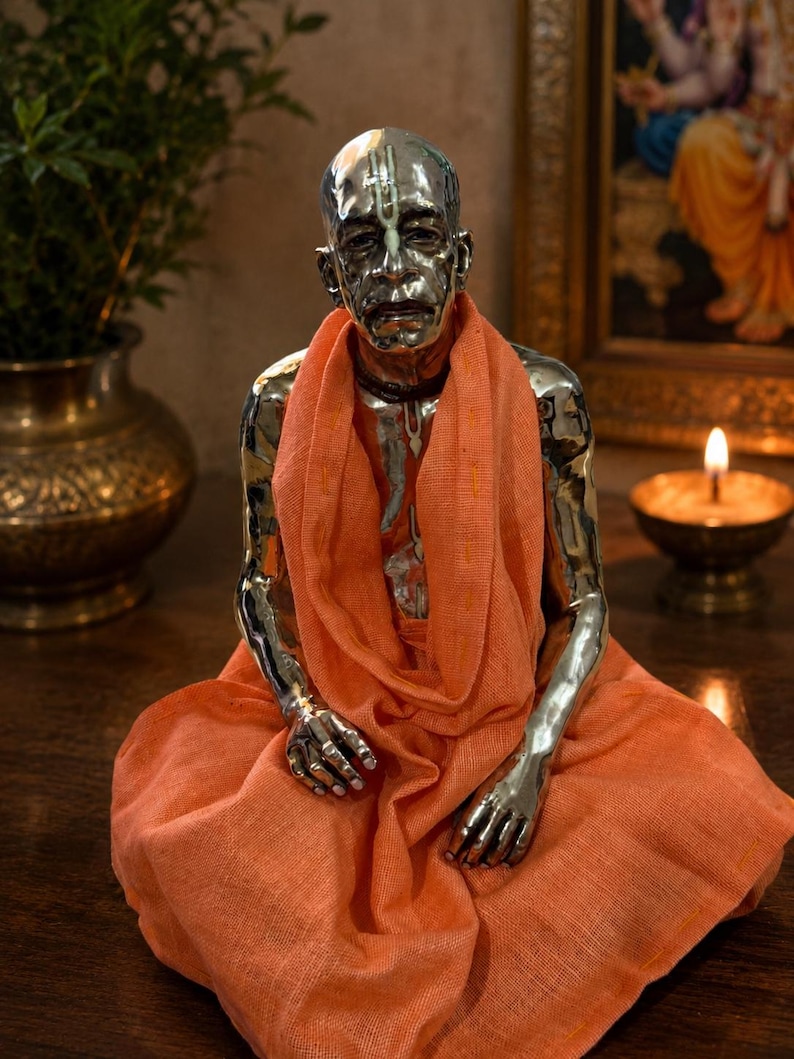 May include: A metallic silver-toned figurine of a seated person with a shaved head, wearing a draped orange fabric garment. The figure features detailed facial expressions and markings on the forehead, presented in a seated, meditative posture.