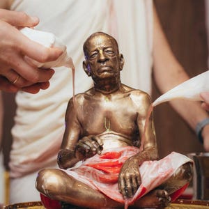 May include: A bronze-colored statue of an elderly person sitting in a cross-legged position on a gold-toned pedestal. The statue is being bathed with a pink liquid poured from white conch shells, with red fabric draped over the lap.
