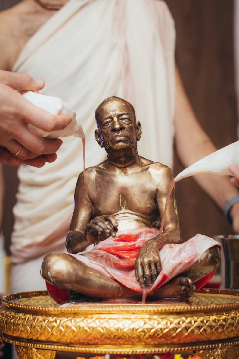 May include: A bronze-colored statue of an elderly person sitting in a cross-legged position on a gold-toned pedestal. The statue is being bathed with a pink liquid poured from white conch shells, with red fabric draped over the lap.