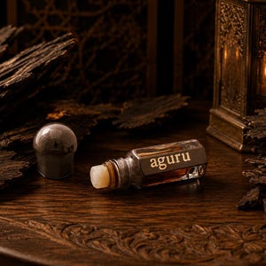 May include: A small glass perfume bottle with a metallic cap and the word aguru printed on the side. The bottle contains a dark amber liquid and is positioned near pieces of dark, textured wood and a metallic lantern with a glowing candle inside.