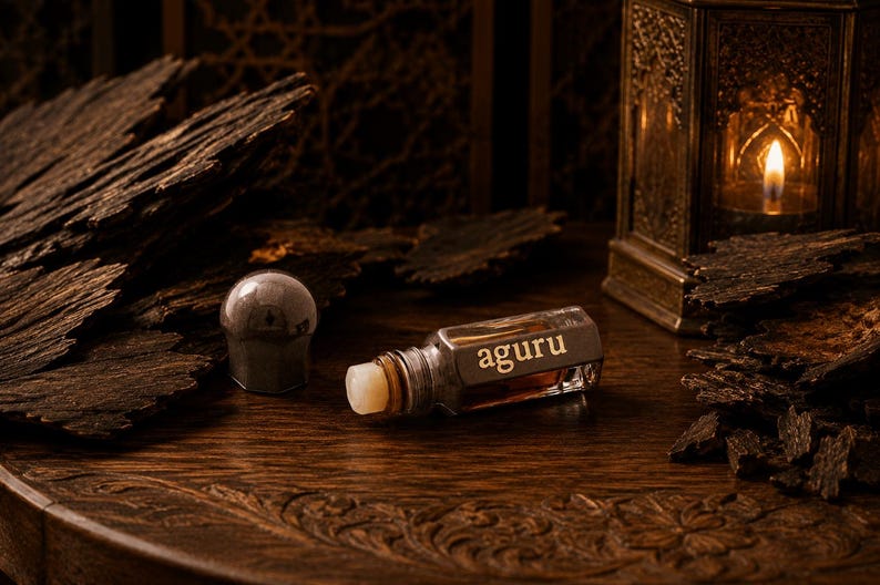 May include: A small glass perfume bottle with a metallic cap and the word aguru printed on the side. The bottle contains a dark amber liquid and is positioned near pieces of dark, textured wood and a metallic lantern with a glowing candle inside.