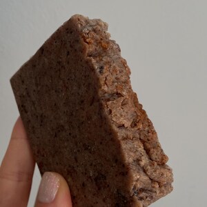 May include: A square, brown food item with a rough, textured edge. The food appears to be a baked good or a type of bread, with a slightly crumbly surface. The color is a mix of brown and reddish hues.