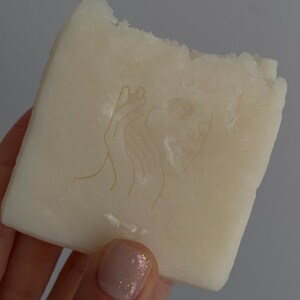 May include: A rectangular bar of off-white soap with a carved design of a woman's face and hand. The soap has a rough, uneven edge, and is held in a hand with a light pink nail polish.