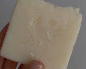 May include: A rectangular bar of off-white soap with a carved design of a woman's face and hand. The soap has a rough, uneven edge, and is held in a hand with a light pink nail polish.
