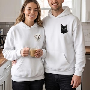May include: Two people wearing matching white hoodies with pocket pouches. One hoodie features a Labrador Retriever graphic, the other a black cat. The couple is standing in a kitchen setting.