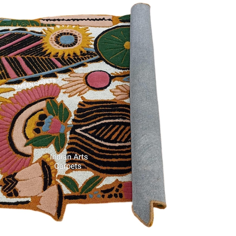 May include: A rolled-up, colorful rug with a floral design. The rug features pink, yellow, black, and green floral patterns on a white background. The text "Indian Arts Carpets" is visible on the rug.