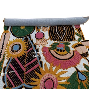 May include: A rolled-up, colorful rug with a floral design. The rug features a white background with large, embroidered flowers in shades of pink, yellow, green, and black. The text "Indian Arts Carpets" is visible on the rug.