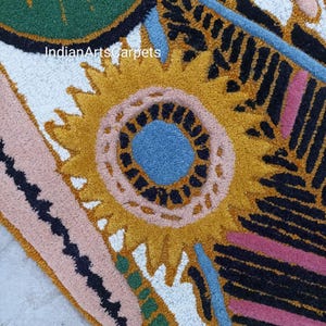 May include: Close-up of a colorful rug with a floral design. The rug features a large yellow sunflower-like shape with a blue center, surrounded by pink, black, and green patterns. The text "IndianArtsCarpets" is visible.