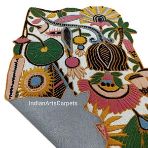 May include: A colorful, floral-patterned rug with a white background. The rug features a variety of flowers and leaves in shades of pink, yellow, green, and blue. The rug's edge is visible, revealing a gray backing. The text "IndianArtsCarpets" is visible.